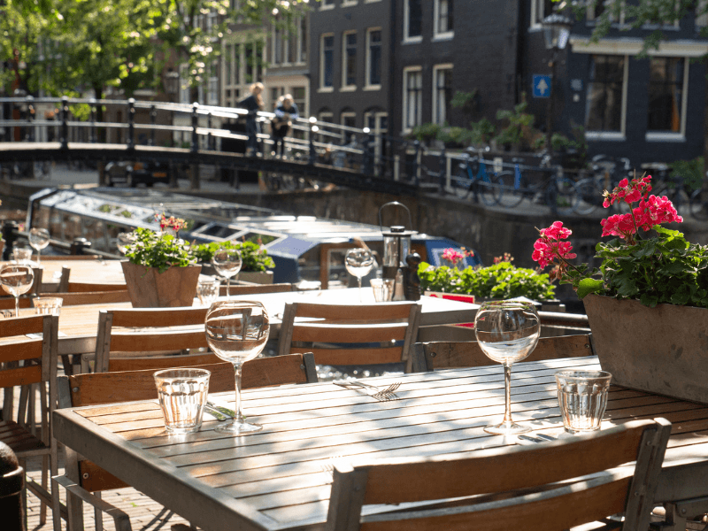 Sonnige Terrasse an einer Amsterdamer Gracht mit Blumen auf dem Tisch, Gläsern und Blick auf eine Brücke und ein Ausflugsboot.